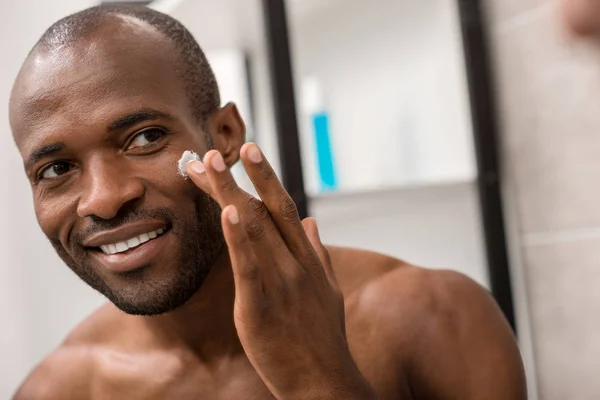 Glücklicher junger Mann, der Gesichtscreme aufträgt, während er im Badezimmer in den Spiegel schaut — Stockfoto