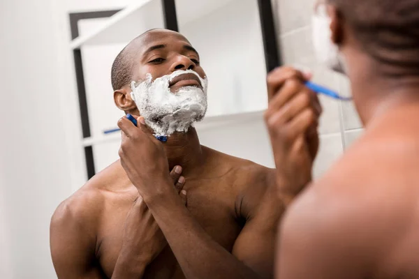 Bel homme afro-américain rasage avec mousse et rasoir et regarder miroir — Photo de stock