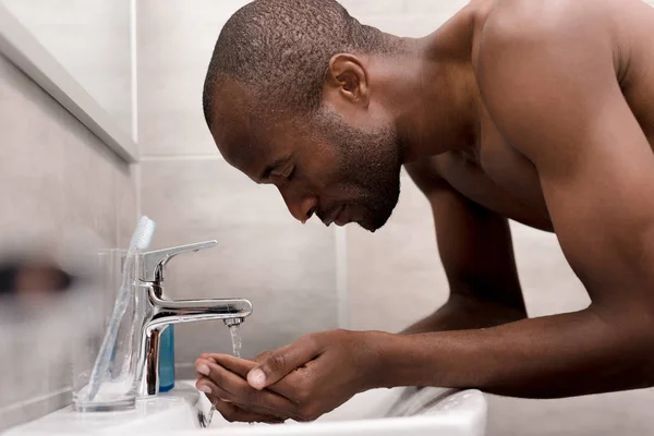 Вид сбоку на молодого небритого афроамериканца, моющего руки в ванной — стоковое фото