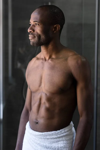 Handsome bare-chested african american man in towel standing and looking away in bathroom — Stock Photo