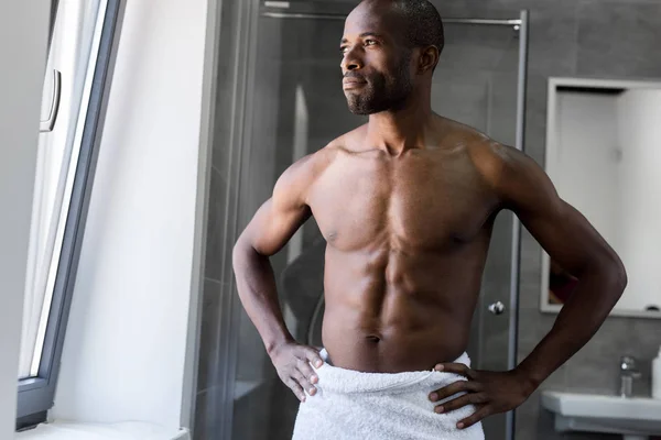 Guapo desnudo pecho africano americano hombre en toalla de pie con las manos en la cintura y mirando hacia otro lado en el baño - foto de stock