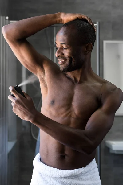 Sorrindo sem camisa afro-americano homem em toalha segurando spray desodorizante no banheiro — Fotografia de Stock