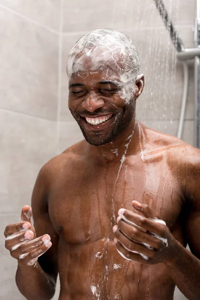 Sorridente jovem afro-americano homem cabeça de lavagem com shampoo no chuveiro — Fotografia de Stock