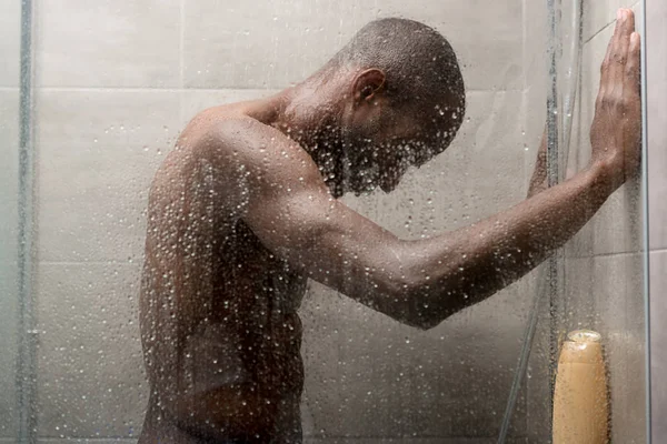 Vista lateral de guapo joven afroamericano hombre lavado cuerpo en ducha - foto de stock