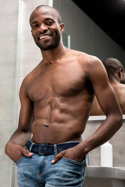 Low angle view of handsome shirtless african american man standing with hands in pockets and smiling at camera in bathroom — Stock Photo