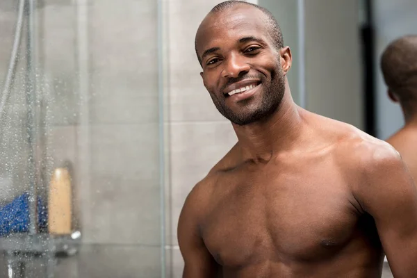 Guapo desnudo pecho africano americano hombre de pie en el baño y sonriendo a la cámara - foto de stock