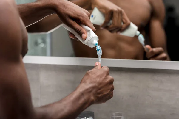 Close-up partial view of young african american man holding toothpaste and toothbrush in bathroom — Stock Photo