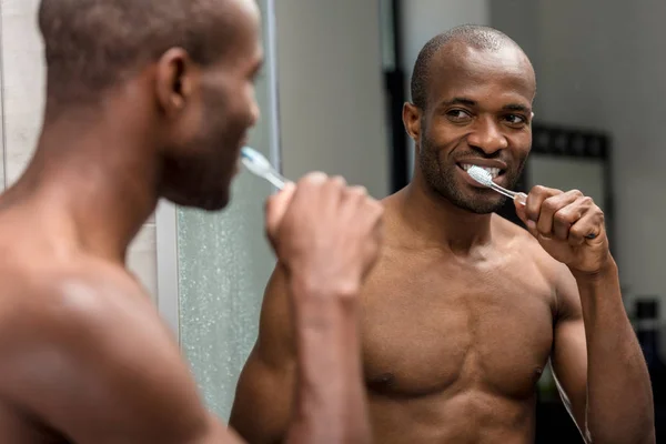 Foco seletivo de sorrir sem camisa homem americano africano escovando dentes e olhando para o espelho — Fotografia de Stock