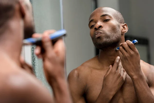 Recortado disparo de joven afroamericano hombre afeitándose con trimmer eléctrico y mirando el espejo - foto de stock