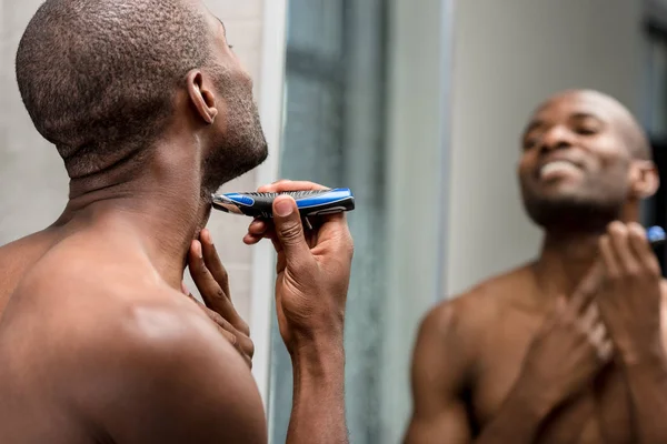 Joven desnudo pecho africano americano hombre afeitándose con trimmer eléctrico en cuarto de baño - foto de stock