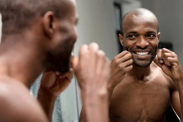Улыбающийся молодой африканский американец держит зубную нить и смотрит в зеркало в ванной комнате — стоковое фото