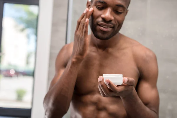 Усміхнений без сорочки молодий афроамериканський чоловік застосовує крем для обличчя у ванній — стокове фото