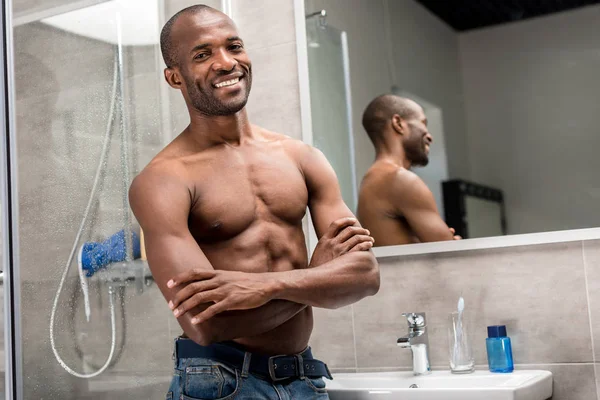 Joven hombre afroamericano con el pecho desnudo de pie con los brazos cruzados y sonriendo a la cámara en el baño - foto de stock