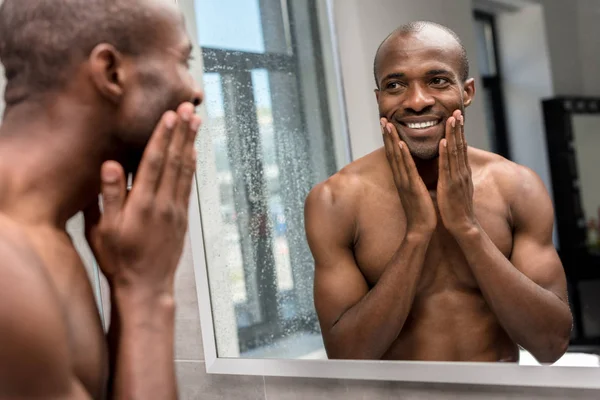 Sorrindo sem camisa afro-americano homem aplicando loção de barbear e olhando para o espelho — Fotografia de Stock