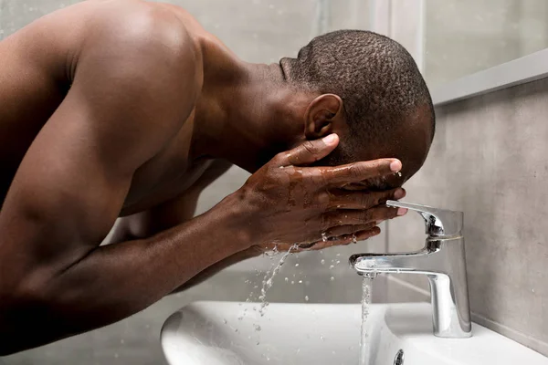 Vista lateral del guapo hombre afroamericano sin camisa lavándose en el baño - foto de stock
