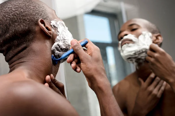 Foco seletivo de belo homem afro-americano barbeando e olhando para o espelho — Fotografia de Stock