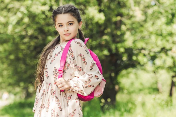 Hermosa colegiala en vestido con mochila mirando a la cámara en el parque - foto de stock