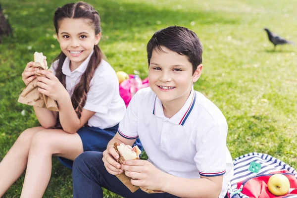 Escolares sonrientes sentados en la hierba y comiendo sándwiches - foto de stock