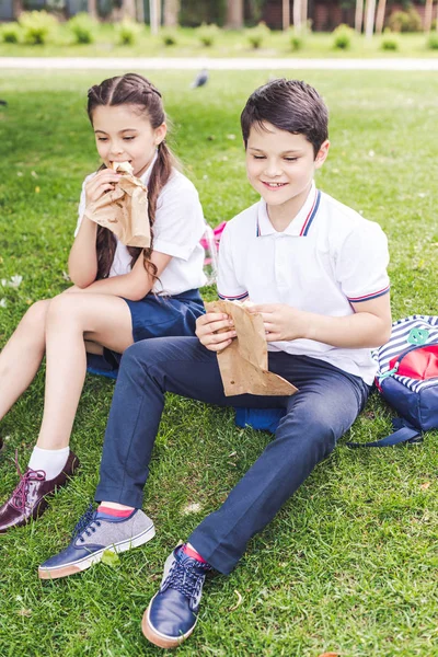 Adorable schoolchildren sitting on grass and eating sandwiches — Stock Photo