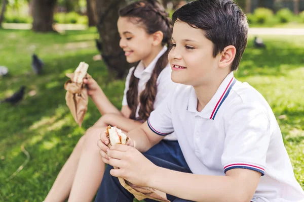 Happy schoolchildren sitting on grass and eating sandwiches — Stock Photo