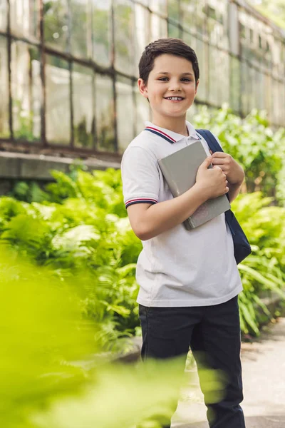 Écolier souriant avec sac à dos et livre regardant la caméra dans le jardin — Photo de stock