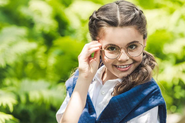 Retrato de cerca de colegiala en camisa blanca y jersey sobre los hombros mirando a la cámara a través de gafas con estilo - foto de stock