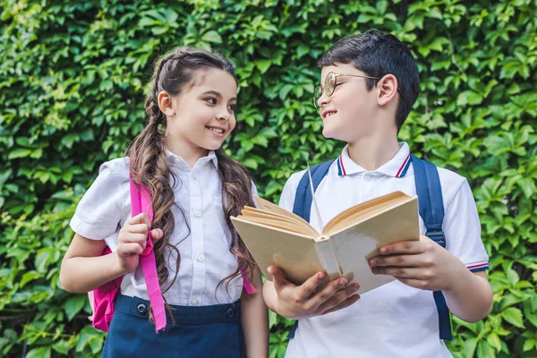 Vista inferior de escolares lendo livro juntos na frente da parede da videira — Stock Photo