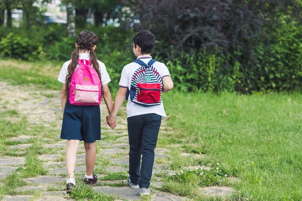 Rückansicht von Schulkindern, die im Park auf dem Gehweg gehen und sich an den Händen halten — Stockfoto