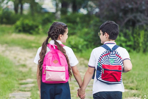 Rückansicht von entzückenden Schulkindern, die im Park auf dem Gehweg gehen und sich an den Händen halten — Stockfoto