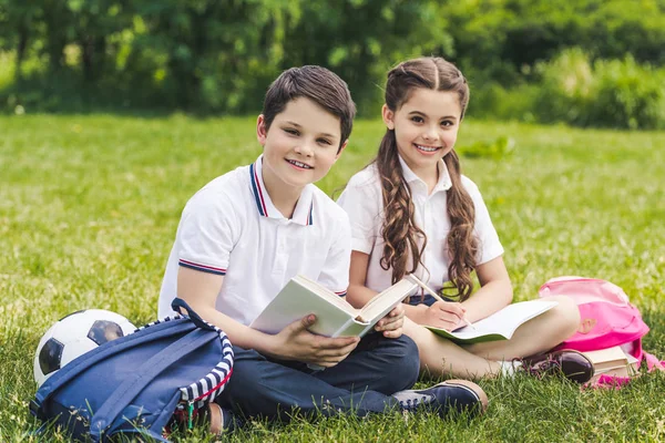 Lächelnde Schulkinder, die gemeinsam Hausaufgaben machen, während sie im Park auf Gras sitzen und in die Kamera schauen — Stockfoto