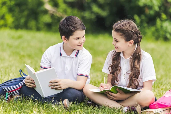 Glückliche Schulkinder, die gemeinsam Hausaufgaben machen, während sie im Park auf Gras sitzen — Stockfoto