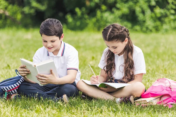 Schulkinder machen gemeinsam Hausaufgaben, während sie im Park auf Gras sitzen — Stockfoto