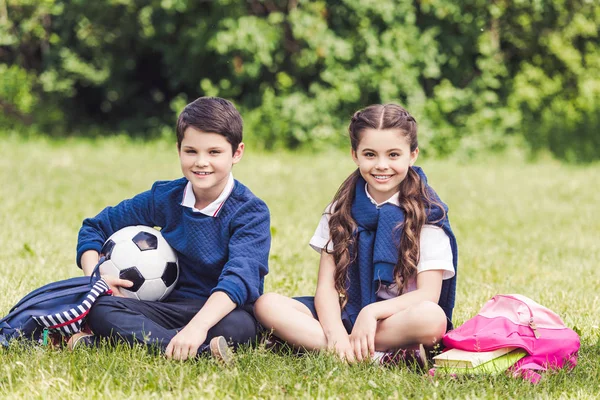 Счастливые школьники сидят на траве в парке с рюкзаками и футбольным мячом — стоковое фото
