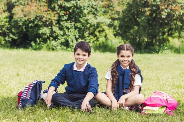 Счастливые школьники, сидящие на траве в парке с рюкзаками — стоковое фото
