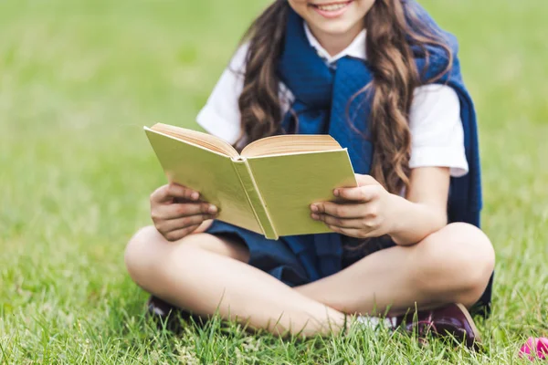 Tiro cortado de colegial sorridente com livro sentado na grama no parque — Stock Photo