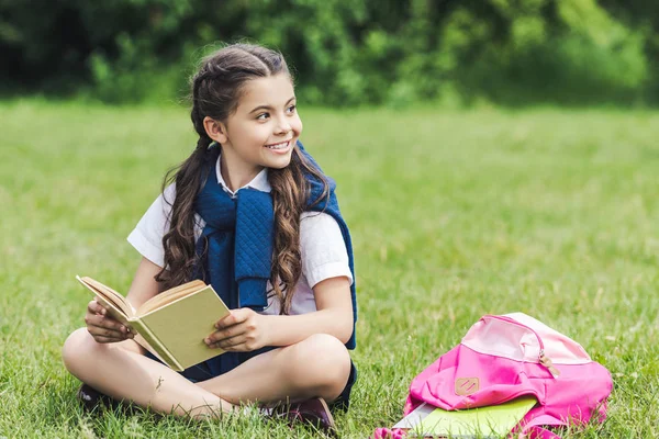 Scolaretta sorridente con libro seduta sull'erba nel parco e distogliendo lo sguardo — Foto stock