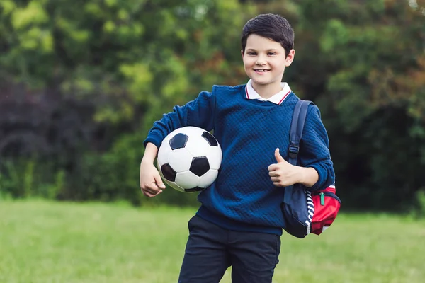 Niño feliz con pelota de fútbol y mochila mostrando el pulgar hacia arriba en el campo de hierba - foto de stock