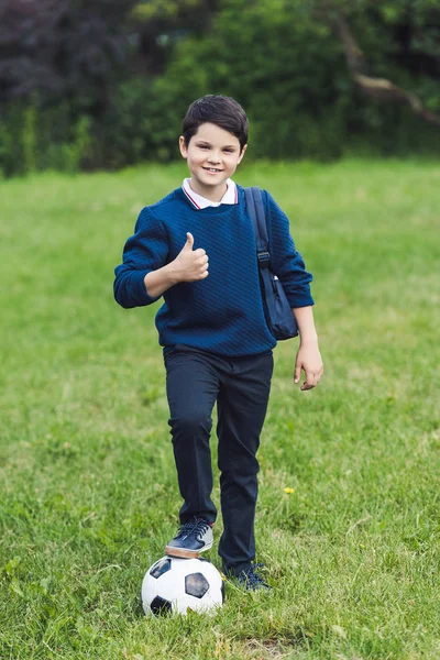 Lächelndes Kind mit Fußball und Rucksack zeigt Daumen hoch auf dem Rasenplatz — Stockfoto