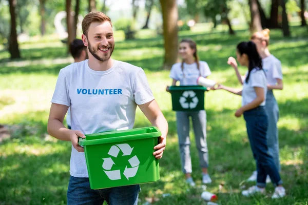 Macho sonriente voluntario celebración reciclaje caja en parque con amigos en fondo - foto de stock