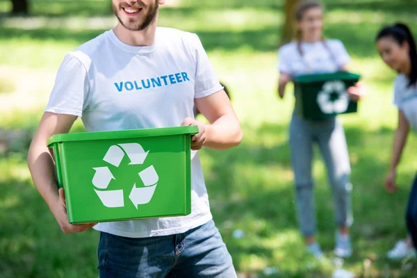 Vista recortada de voluntarios sosteniendo caja de reciclaje verde en el parque con amigos en el fondo - foto de stock