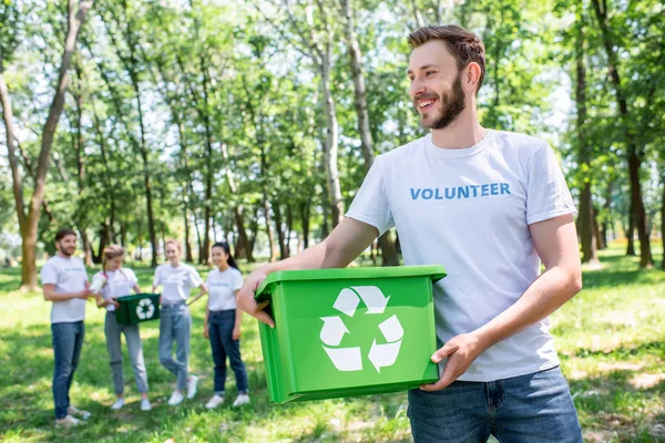 Joven voluntario sosteniendo caja de reciclaje en el parque con amigos en el fondo - foto de stock