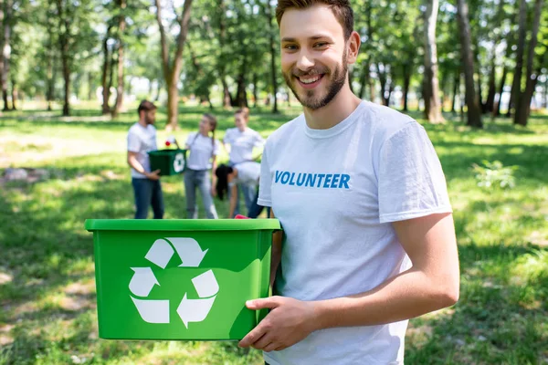 Joven voluntario sonriente con caja de reciclaje verde en el parque con amigos en el fondo - foto de stock