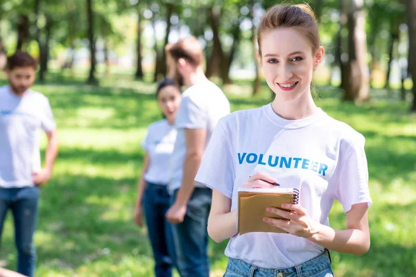 Female volunteer writing in textbook while people cleaning park — Stock Photo
