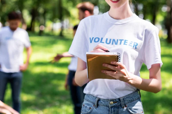 Девушка пишет в учебнике, пока волонтеры убирают зеленый парк — стоковое фото