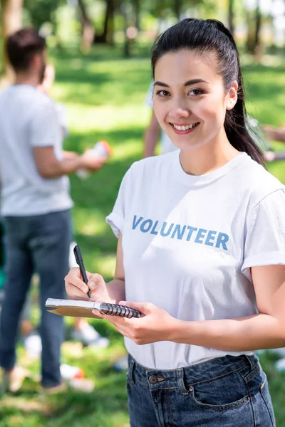 Voluntario escribiendo en un libro de texto mientras los amigos limpian el parque - foto de stock