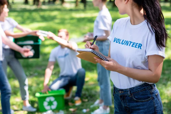 Mujer escribiendo en libro de texto mientras voluntarios limpian el parque - foto de stock