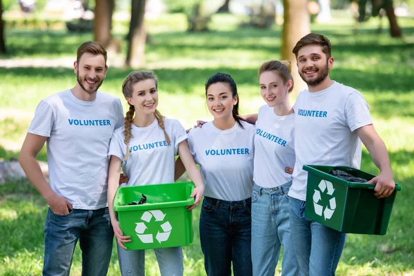 Jóvenes voluntarios con cajas de reciclaje verdes para la basura de pie en el parque - foto de stock