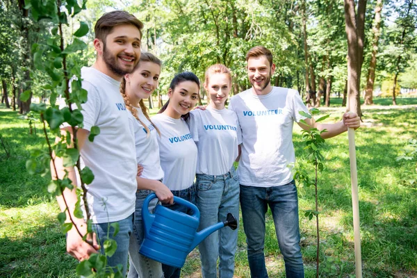 Voluntarios con regadera y árboles nuevos de pie en el parque juntos - foto de stock