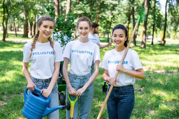 Young female volunteers with watering can, shovel and rake standing in park — Stock Photo