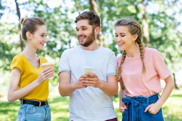 Amis souriants avec du café pour aller utiliser le smartphone ensemble dans le parc — Photo de stock